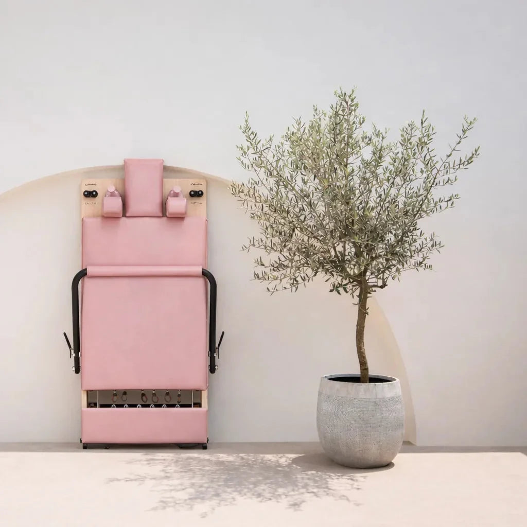 Machine reformer Pilates pliable couleur rose rangée verticalement contre un mur blanc avec olivier en pot - Pilates At Home