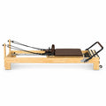 Reformer Pilates studio en bois clair avec carrosserie marron, sangles et barre de pieds, vue de profil - Pilates At Home