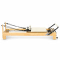Reformer Pilates studio en bois clair avec carrossage en tissu beige, câbles et repose-pieds, vue de profil - Pilates At Home